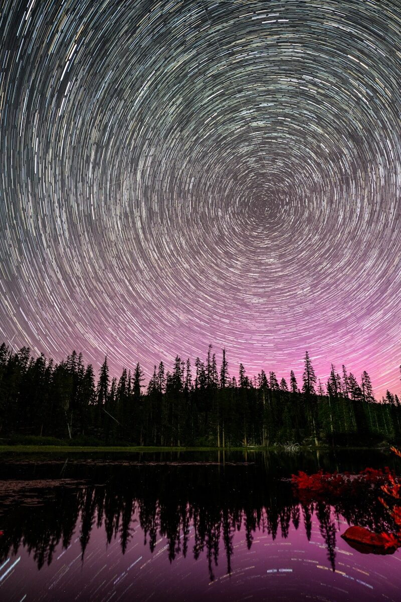 Star trails over forest and lake with purple sky.